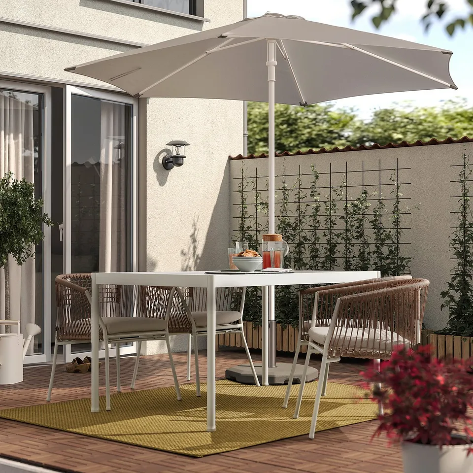 Outdoor patio with white MORUM rug, wicker chairs, white dining table, and large beige umbrella on wooden deck, plant in foreground.