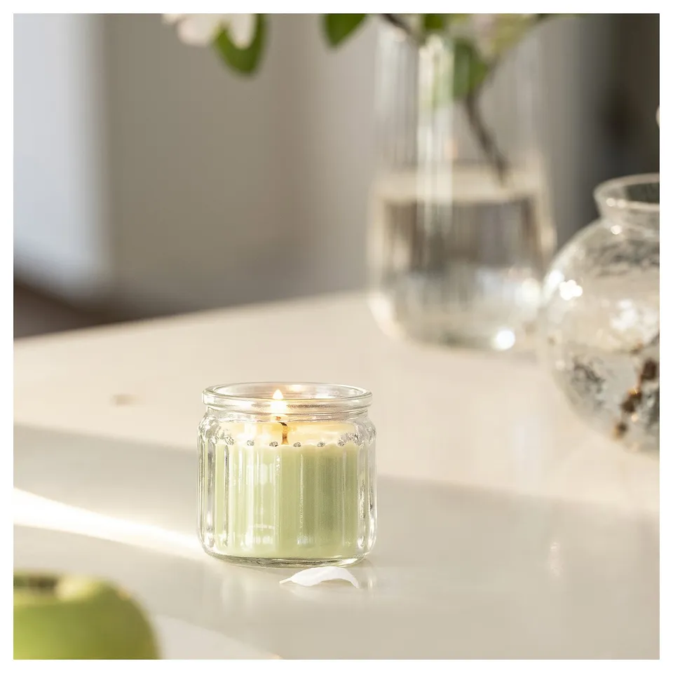 A lit apple-scented candle in a clear glass jar sits on a white countertop beside two empty vases.