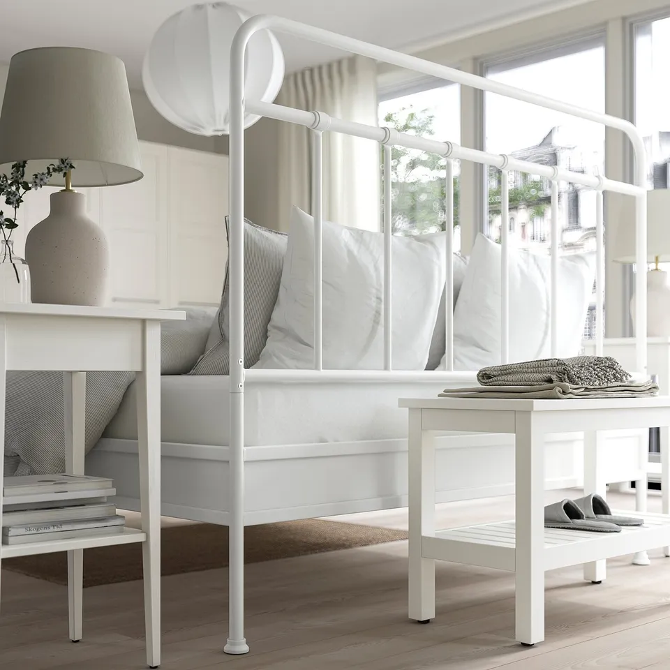 Minimalist bedroom with white metal bed, grey bedding, lamp, vase, and window view.