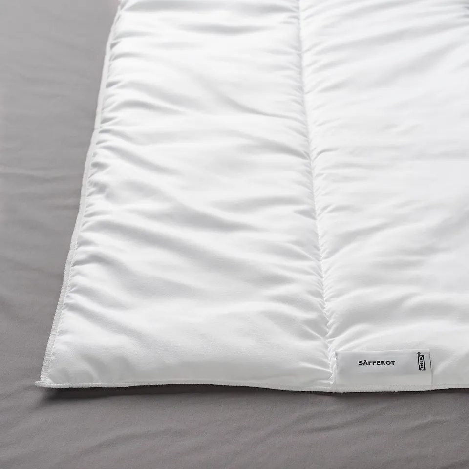 White duvet on a grey bedsheet, showing smooth texture and feather-like stitching, with a tag reading SÄFFEROT near the corner.
