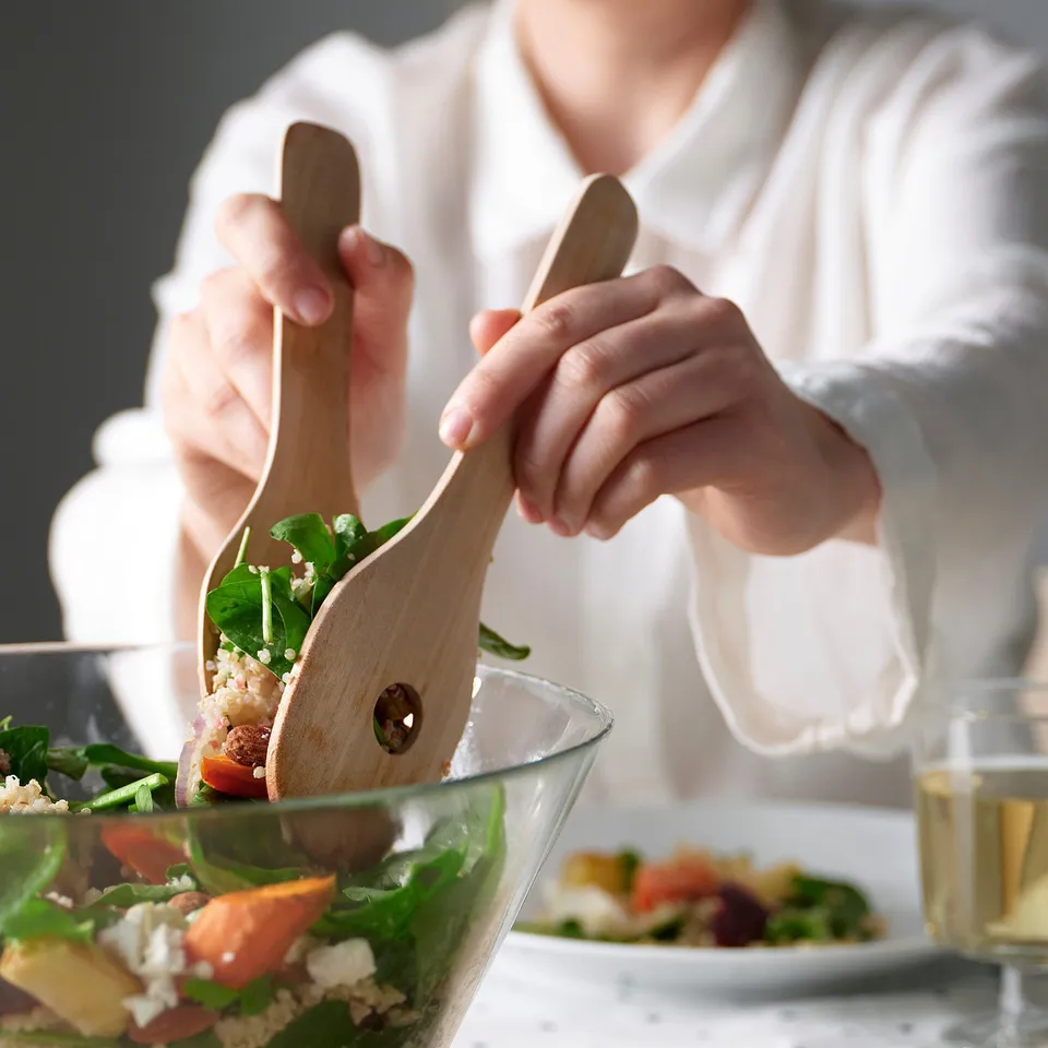 Person using wooden salad servers to toss a mixed salad in a glass bowl. The servers have a simple, rounded design for comfortable handling.