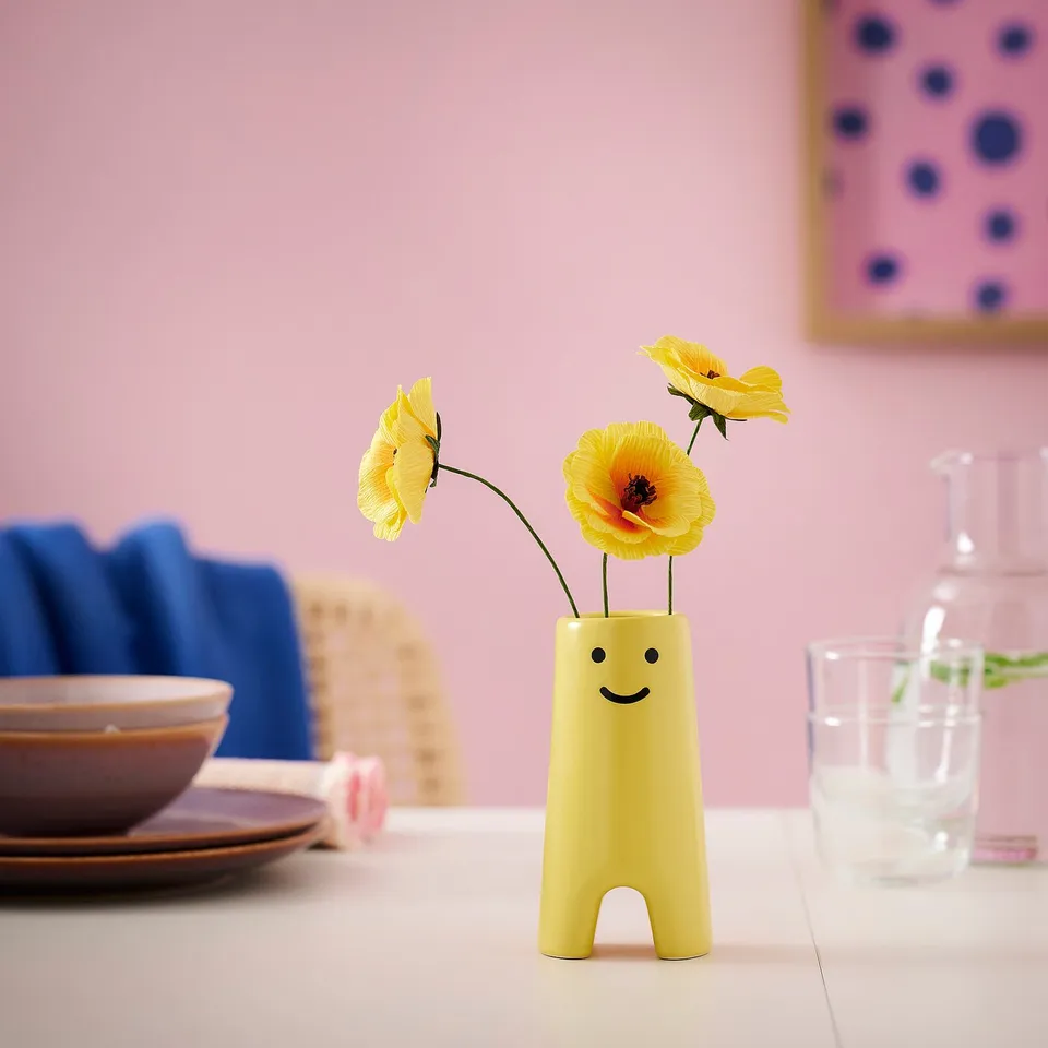 Small yellow FRÖVÄXT vase with smiley face, made of stoneware, holding yellow flowers, sitting on a table with dishes and a glass.