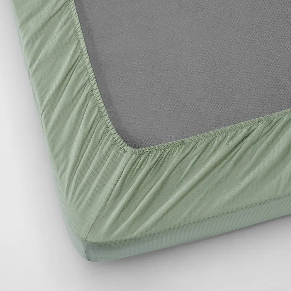 A green fitted sheet with a soft pattern, sized for mattresses up to 26 cm thick, featuring elastic edging for a secure fit.
