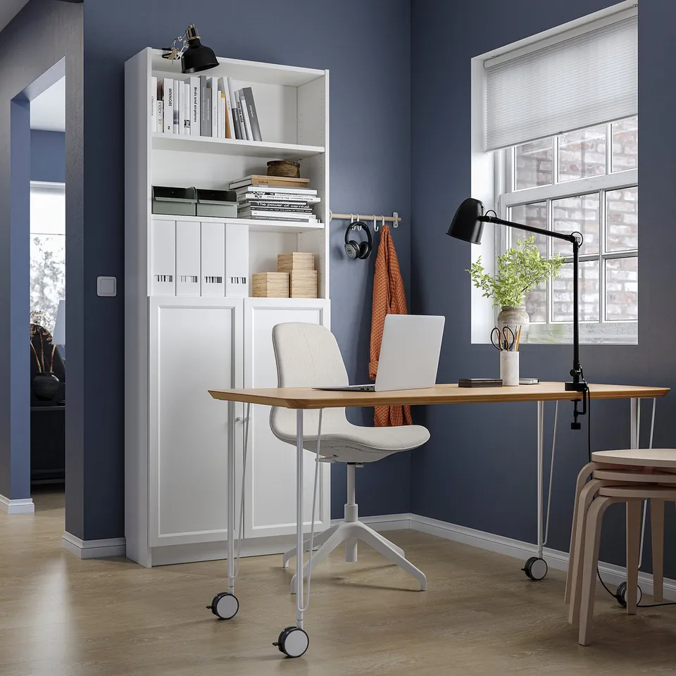 Home office with white BILLY bookcase, wooden desk, grey chair, black lamp, window with blinds, and plant.