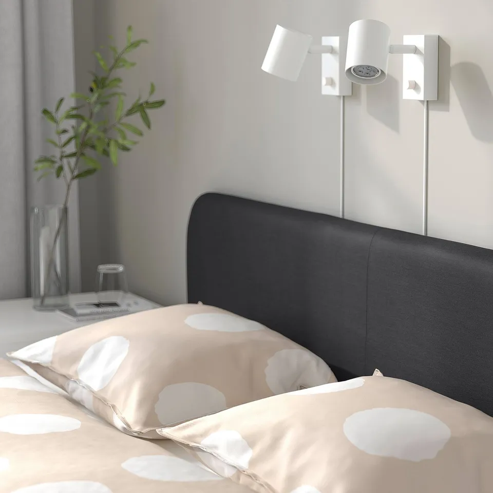 Bedroom with black upholstered bed, light beige bedding, wall-mounted white reading light. Plant and glass vase on bedside table.
