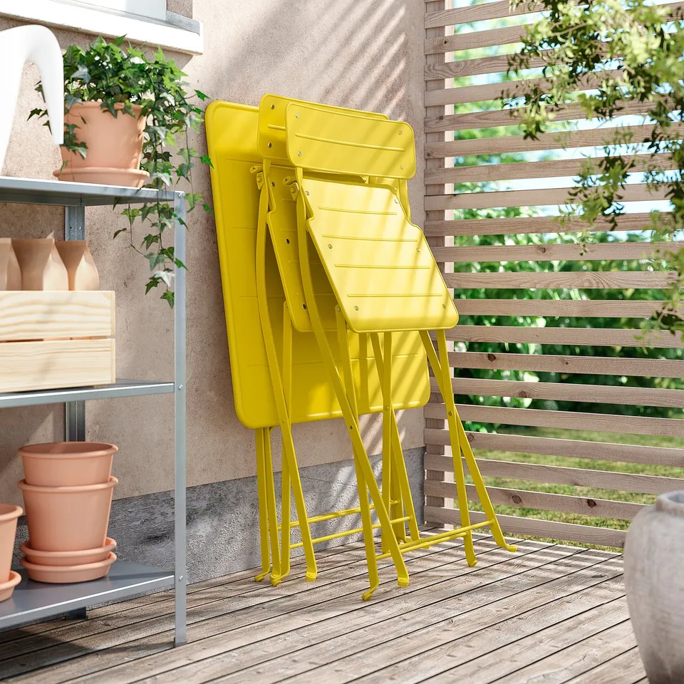 Bright yellow SUNDSÖ folding chair, stacked on balcony, wooden deck. Made of durable steel, perfect for small spaces.