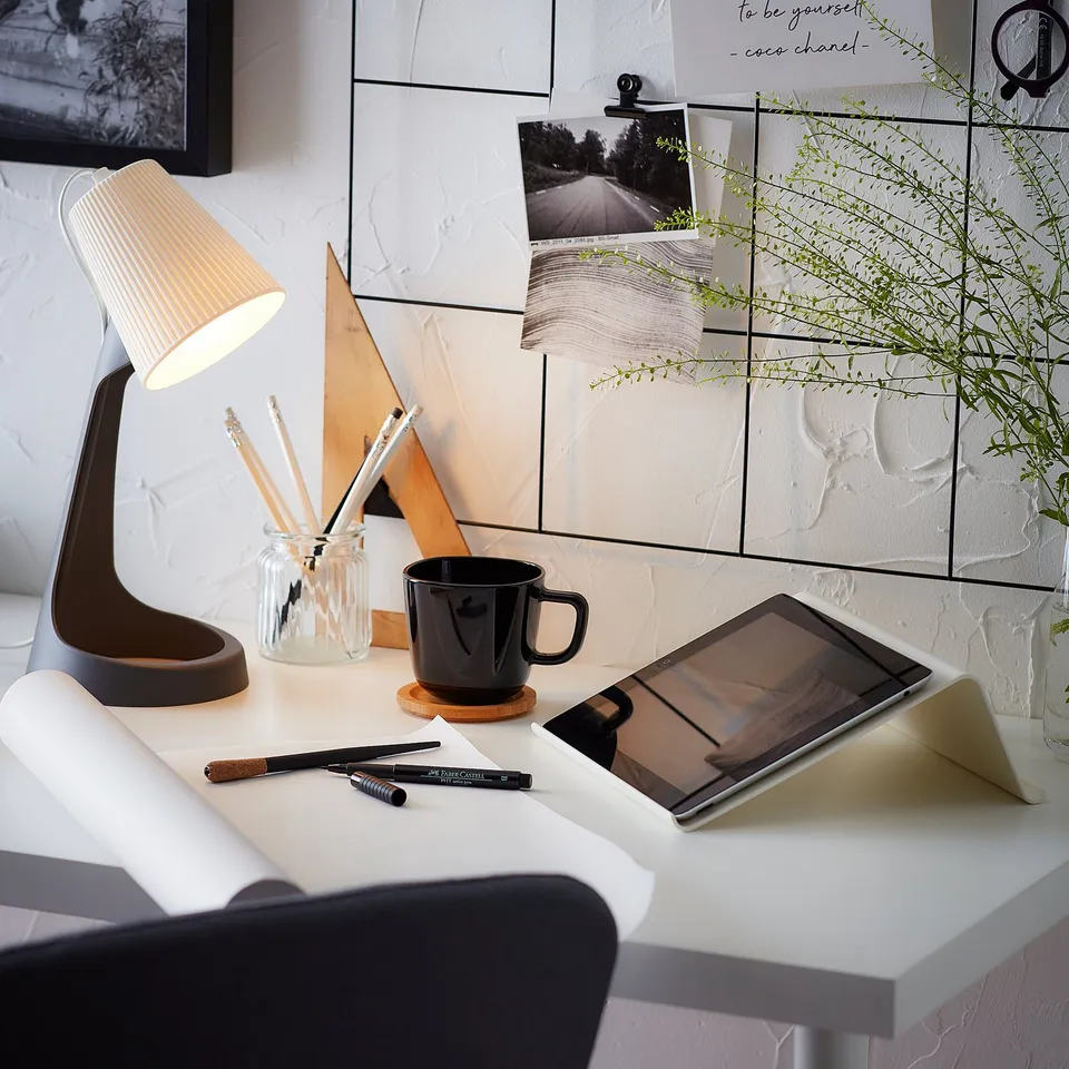 A minimalist workspace featuring a white desk with a lamp, tablet, and mug. A mirror with photos and plants decorates the wall.