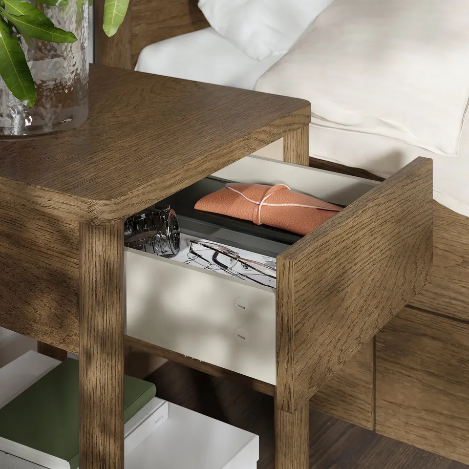 Wooden bedside table with open drawer revealing orange cloth, beside a bed with white sheets.
