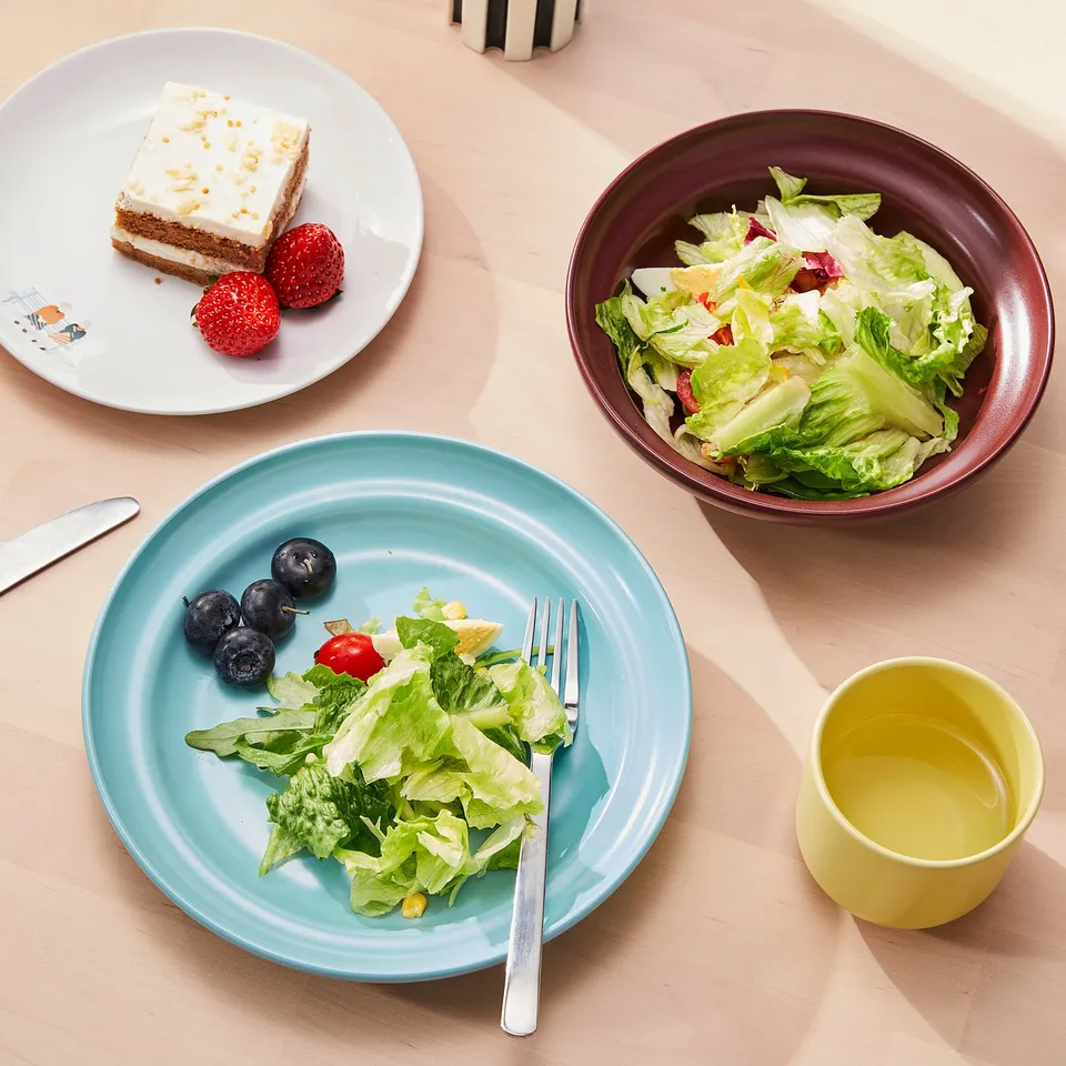 Three plates with food: salad, cake, and berries, plus a bowl, fork, and yellow cup on a table, suggesting a meal setting.