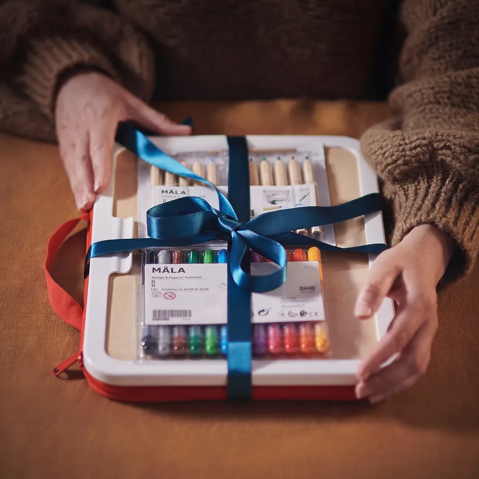 A gift-wrapped MÅLA drawing case in white, tied with blue ribbon, containing art supplies.