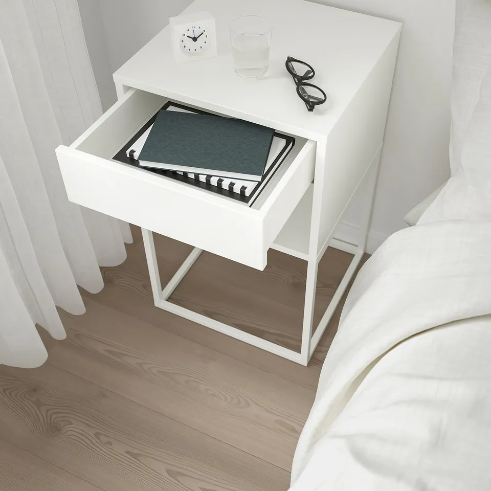 White bedside table with open drawer showing book, next to bed with white sheets.