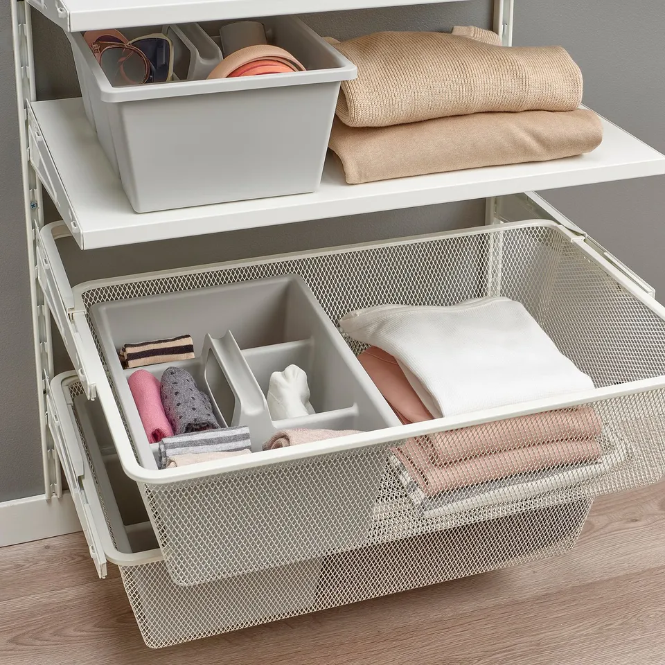 White wall-mounted storage unit with shelves and drawers, holding folded clothes, accessories in boxes, and rolled items in a mesh drawer.