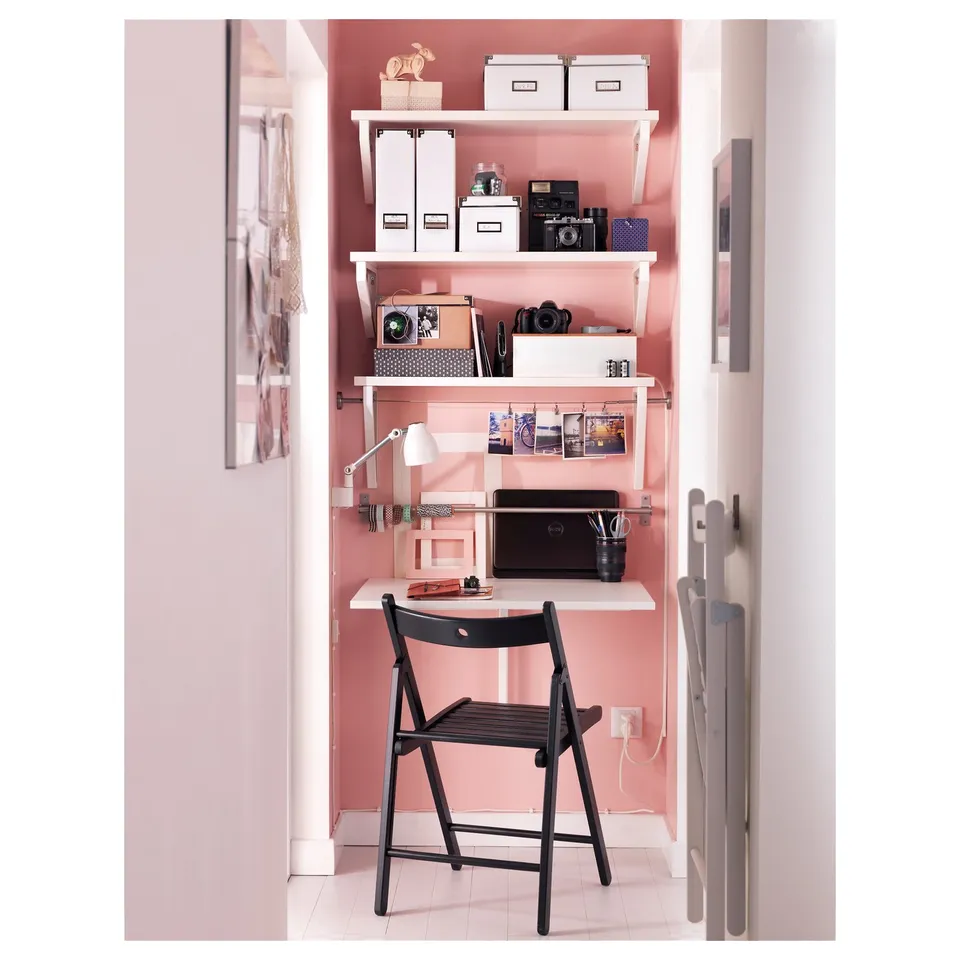 A compact home office setup with a black folding chair and open shelving in a pink alcove.