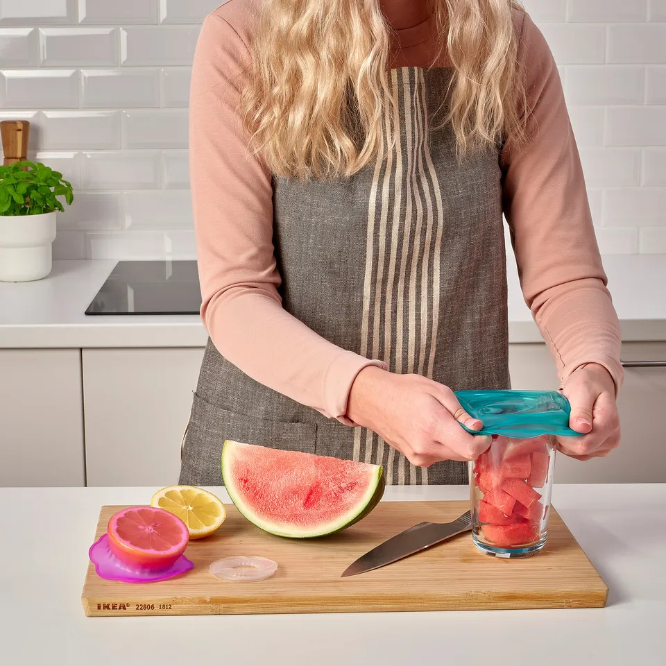 Person preparing fruits with overmatt food covers. Shes covering a jar with watermelon and using a lemon, grapefruit, and knife on a cutting board.