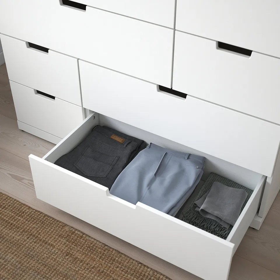 An open drawer of a white NORDLI chest, holding neatly folded pants. The product offers modern, practical storage.