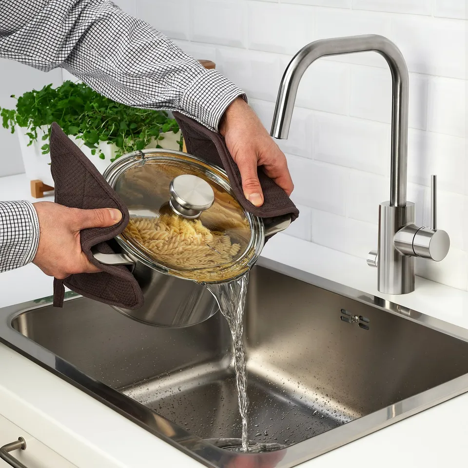 Person pours boiled pasta into stainless steel sink with modern tap.