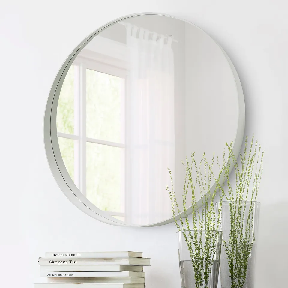 A round mirror hangs on a white wall reflecting a window. Below it, a stack of books and a vase with green plants are visible.