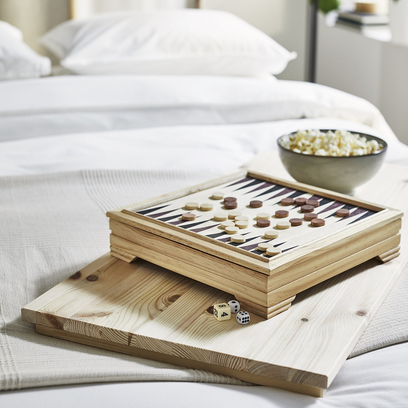Bedside setup with GLAMBERGET extendable bed, wooden backgammon game, popcorn bowl, and dice on a tray.