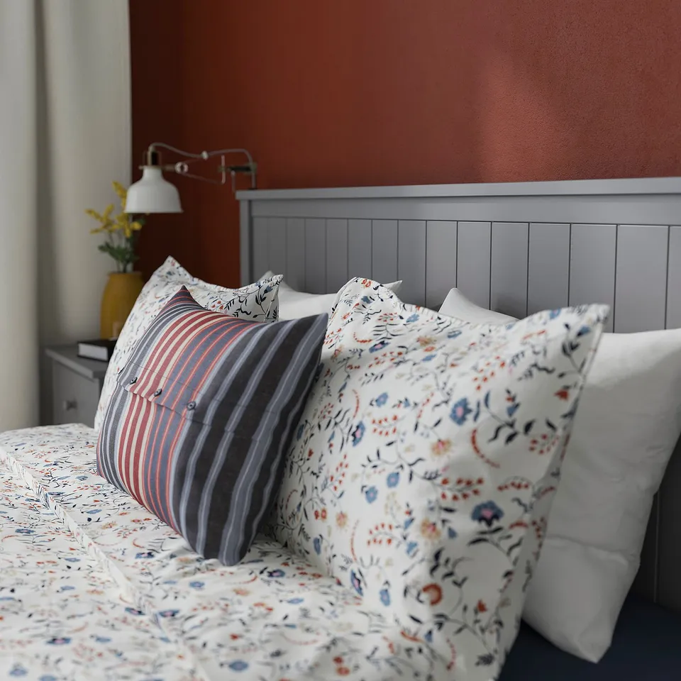 Cosy bed with floral patterned pillows and striped cushion against a wooden headboard, set against red wall with a wall lamp and vase.