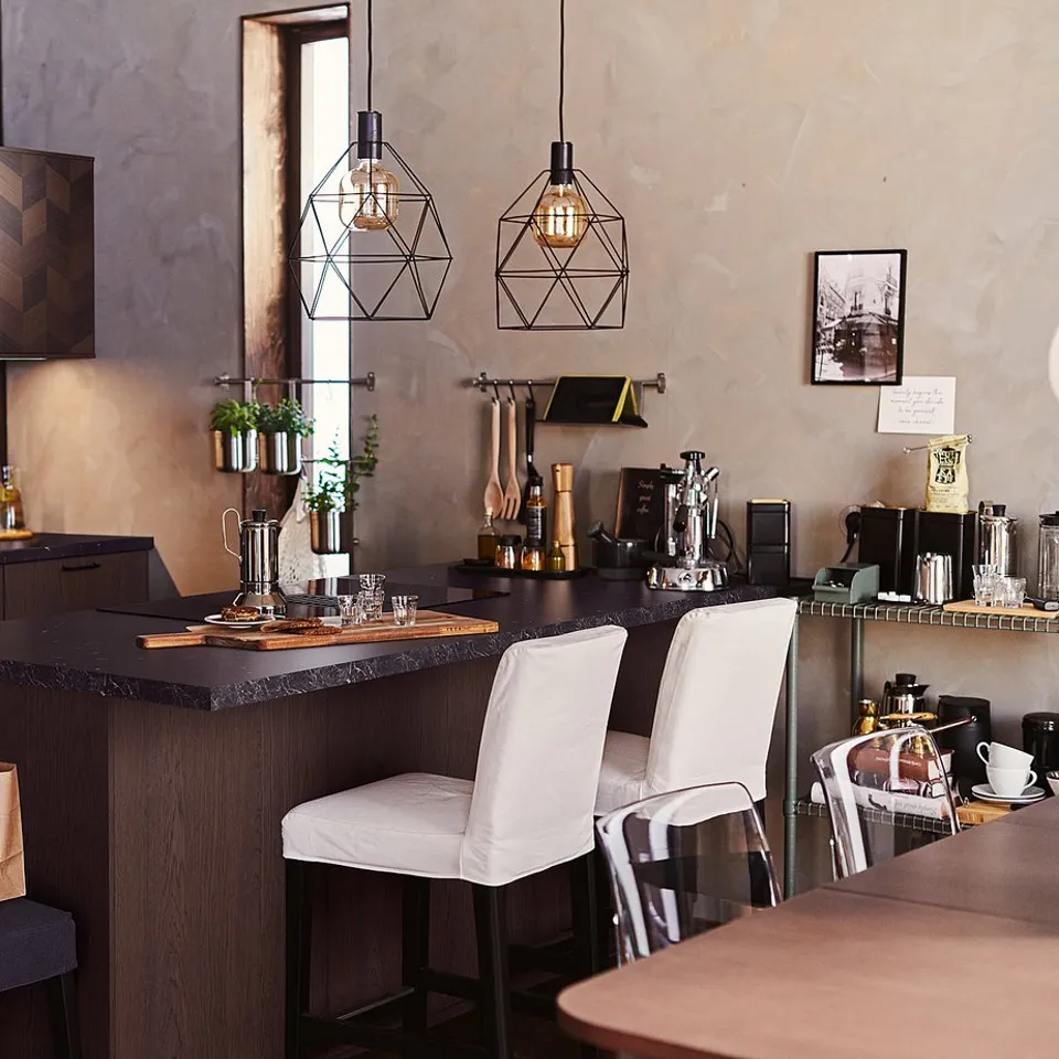 Modern kitchen with dark wood cabinets, geometric pendant lights, sleek countertop, white chairs, hanging utensils, and decorative items.