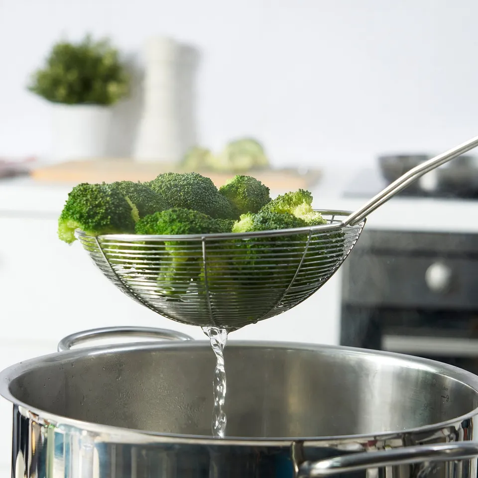 Stainless steel skimmer removing broccoli from pot, showcasing its ability to drain liquid efficiently.