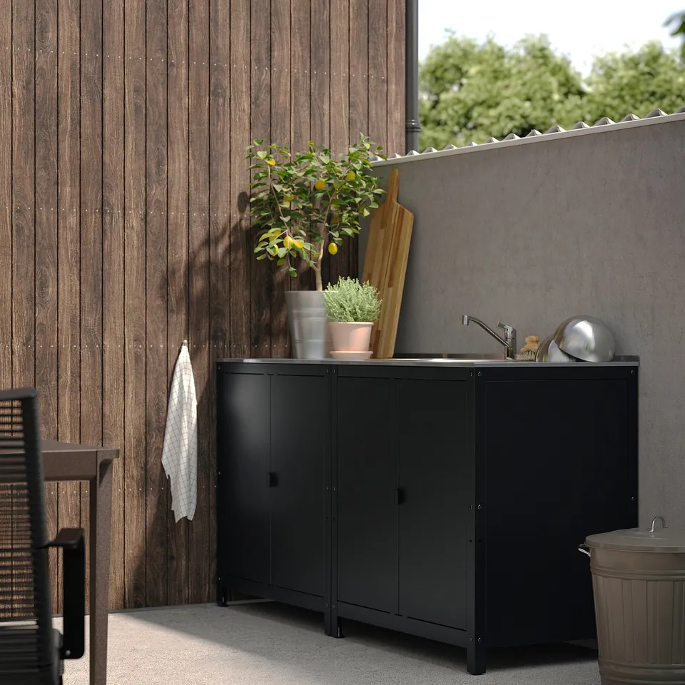 Modern outdoor black kitchen unit with sink, shelves, and plants. Fits neatly against wooden fence, smaller than adjacent bin.
