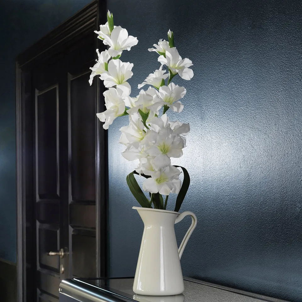White enamel vase on shelf, containing tall white flowers, set against blue wall.