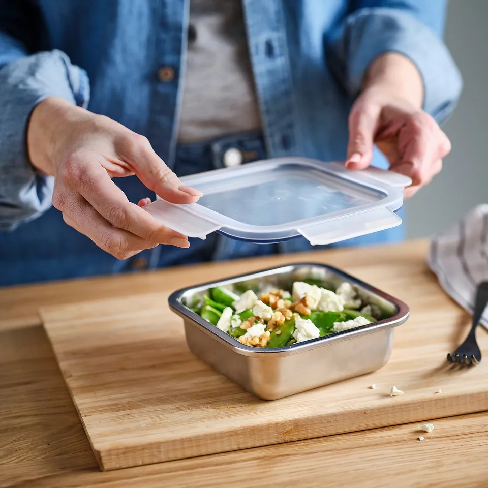 Person opens food container lid, revealing salad with feta and nuts inside. The container sits on a wooden table, ready for meal prep or storage.