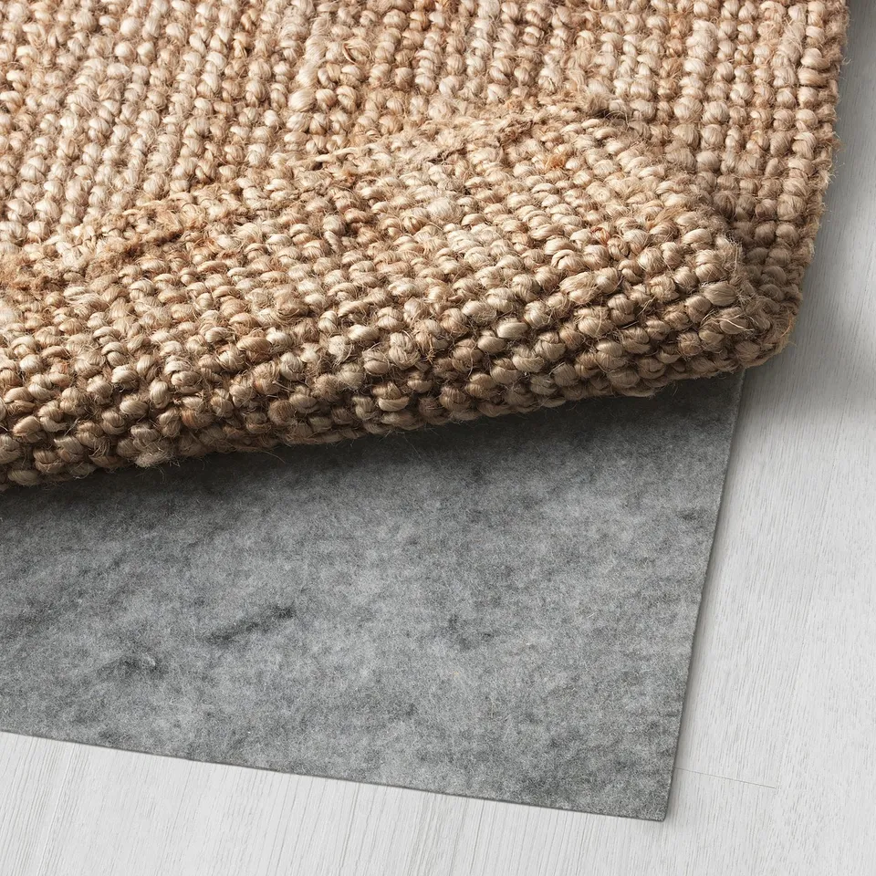 Light brown, hand-woven rug laid over dark grey rug underlay, showing its textured surface and thickness.