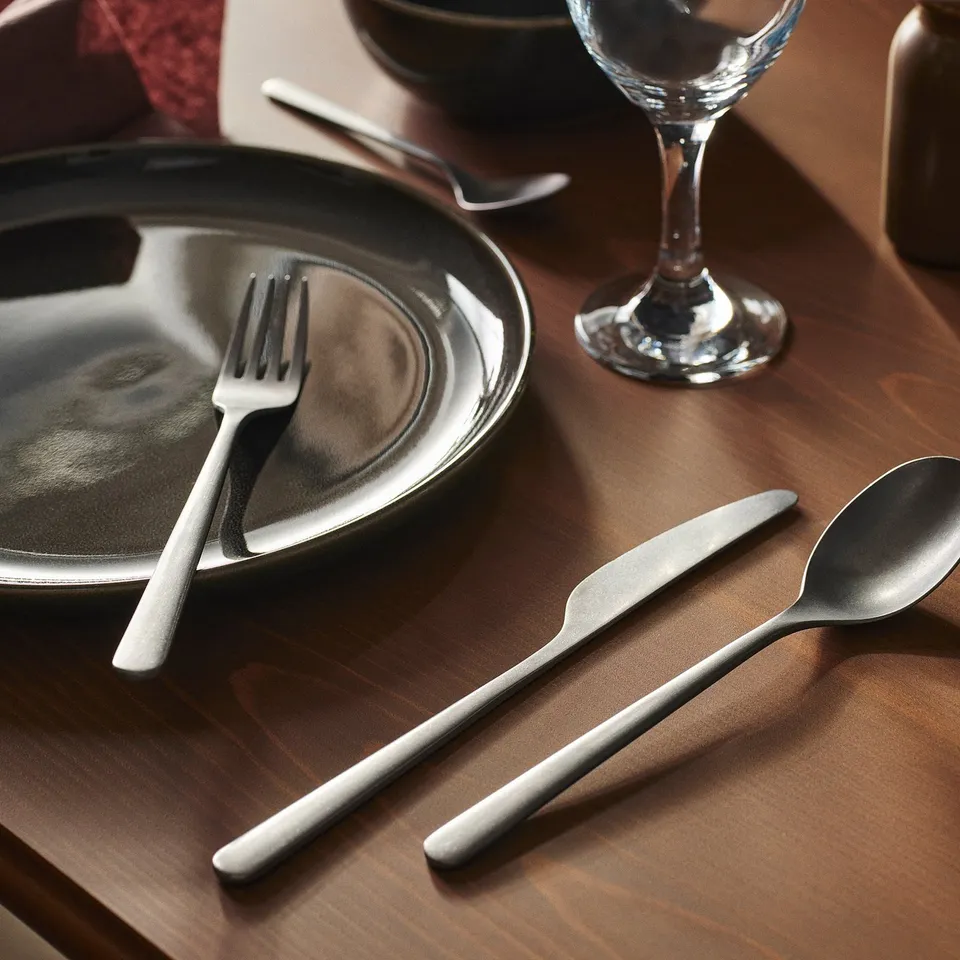 Silver cutlery set on wooden table with dark plate and wine glass.